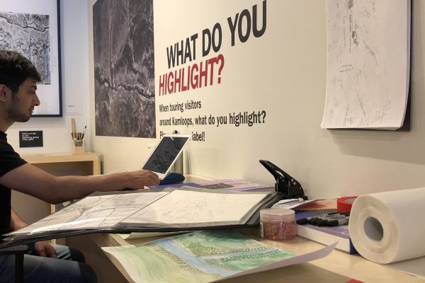 Maps and pieces of paper in the foreground on a table. Text on wall says 'What do you highlight?" in relation to an aerial map of Kamloops. A seated man is working on a laptop at the right of the photo.